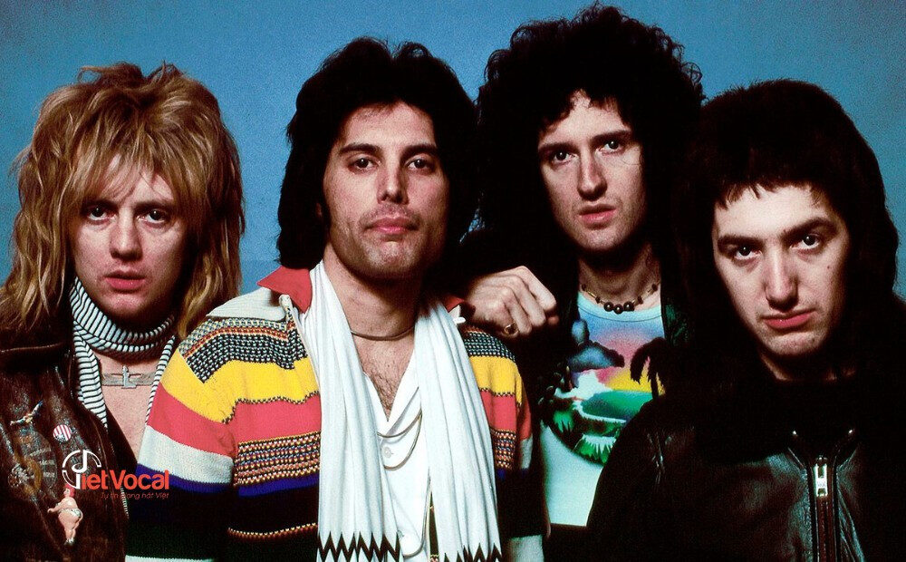 alt text: Ban nhạc Queen biểu diễn trên sân khấu, với Freddie Mercury hát chính, Brian May chơi guitar, John Deacon chơi bass và Roger Taylor chơi trống.
