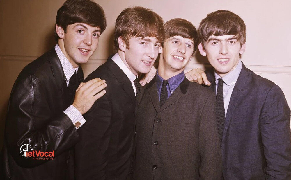 alt text: Ban nhạc The Beatles biểu diễn trên sân khấu, với John Lennon chơi guitar, Paul McCartney hát, George Harrison chơi guitar và Ringo Starr chơi trống.