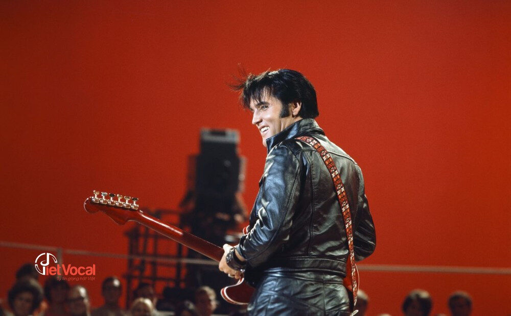alt text: Elvis Presley trong trang phục biểu diễn, đang hát và chơi guitar.